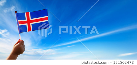 A child's hand holds a small Icelandic flag against a blue sky. 131502478