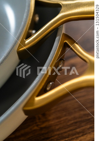 Close-up of curved matte gold handles on nested ceramic pans. The cookware rests on a wood background with a focus on smooth curves and modern design. Close-up of curved matte gold handles on nested ceramic pans. The cookware rests on a wood background with a focus on smooth curves and modern design. 131504329