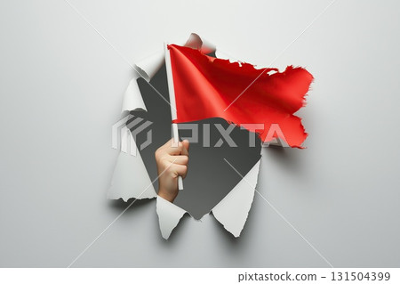 A hand holding a torn red flag emerges through a ripped hole in gray paper 131504399