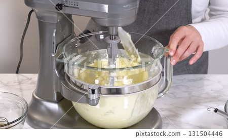 A stand mixer holds creamy cookie dough in a glass bowl set on a marble countertop. The bright light highlights the smooth mixture in a clean kitchen environment. 131504446