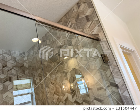 A modern bathroom shower with a frameless glass door and decorative geometric tiles. The lighting emphasizes the texture and layout of the tiled surface. A modern bathroom shower with a frameless glass door and decorative geometric tiles. The lighting emphasizes the texture and layout of the tiled surface. 131504447