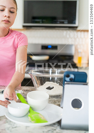 A young girl in a pink t-shirt washes her hands at the kitchen sink. Baking tools and a phone stand are visible on the counter nearby. A young girl in a pink t-shirt washes her hands at the kitchen sink. Baking tools and a phone stand are visible on the counter nearby. 131504469