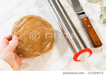 Straight-on view of hands holding amber gingerbread cookie dough wrapped in plastic wrap, showing preparation for chilling. 131504472