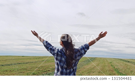 Young woman is embracing the beauty of landscape while raising her arms in the air, feeling the freedom and awe of nature. woman in field with her arms raised up to sky. Young woman is embracing the beauty of landscape while raising her arms in the air, feeling the freedom and awe of nature. woman in field with her arms raised up to sky. 131504481