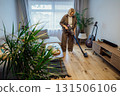 Middle aged woman is vacuuming floor of modern apartment living room with green plants, using portable cordless vacuum cleaner. Daily routine and household chores. Housekeeping, home cleaning concept. 131506106