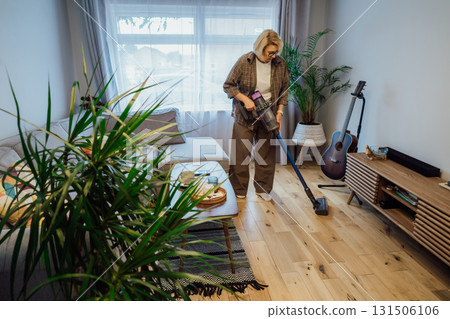 Middle aged woman is vacuuming floor of modern apartment living room with green plants, using portable cordless vacuum cleaner. Daily routine and household chores. Housekeeping, home cleaning concept. 131506106