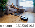 Middle aged woman relaxing on sofa while robot vacuum cleaner doing housework. Smart tech urban house IOT appliances better life. Lady enjoys home care to robot hoover clean floor through mobile app 131506108