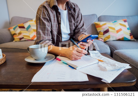 Middle aged woman reviewing expenses, managing personal finances on smartphone mobile app while sitting on couch with coffee in cozy living space. finance planning, financial balance check, budgeting 131506121