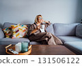 Middle aged smiling woman sitting on sofa and looking away while drinking coffee or tea. Mature senior woman relaxing after housekeeping, home cleaning. Portrait of relaxed female resting at home. 131506122