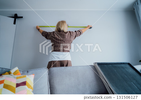 Back view Middle aged woman doing measuring with a measure tape on the wall. Lady wants to put a picture on the wall at home. Housekeeping work. Doing repair herself. DIY, equality in work concept. 131506127