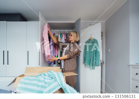 Middle aged woman selecting clothes from wardrobe for donating to Charity shop. Decluttering, Sorting clothes and Cleaning Up. Reuse, second-hand concept. Conscious consumer, sustainable lifestyle. 131506131