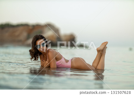 young woman relaxes in the shallow water at the beach. She wears stylish sunglasses and a pink swimsuit enjoying the tranquil atmosphere as the sun sets. young woman relaxes in the shallow water at the beach. She wears stylish sunglasses and a pink swimsuit enjoying the tranquil atmosphere as the sun sets. 131507628