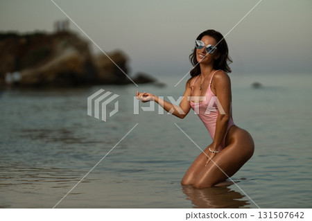 beautiful woman is kneeling in shallow water near the seashore during sunset. She is wearing sunglasses and a swimsuit radiating joy and tranquility amidst gentle waves. 131507642