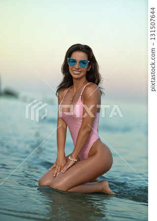 woman kneels in shallow water by the seashore during sunset. She wears a pink swimsuit and blue sunglasses radiating joy and beauty as waves lap at her skin. woman kneels in shallow water by the seashore during sunset. She wears a pink swimsuit and blue sunglasses radiating joy and beauty as waves lap at her skin. 131507644