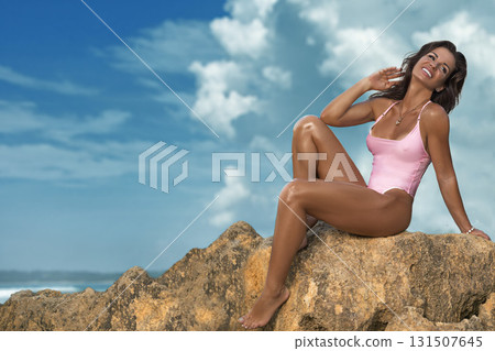 beautiful woman sits on a large rock by the seashore wearing a pink swimsuit. She smiles happily while enjoying the sunny weather and the refreshing ocean breeze. 131507645