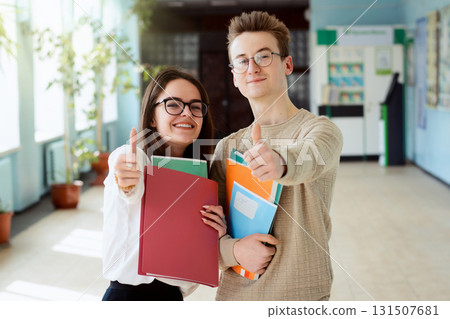 Two happy students in good mood showing thumbs up to camera standing in campus of university, holding books, exercise books, course books. Young learners are eager to study 131507681