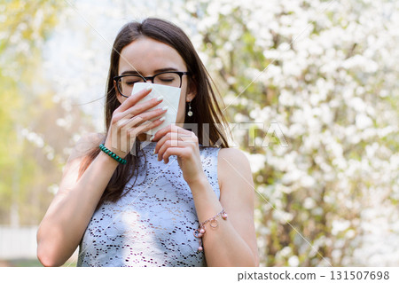 Outdoor shot of discontent young girl with seasonal allergy, uses white tissue, poses over blooming tree, has rhinitis and sneezing, reacts on allergens. People and disease concept 131507698