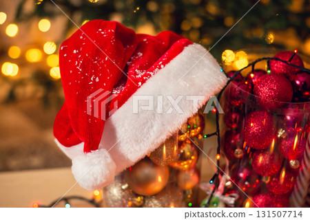 Red christmas hat on a package of golden baubles for fir-tree, close-up. Red balls and spruce beauty with warm lights on the background. Red christmas hat on a package of golden baubles for fir-tree, close-up. Red balls and spruce beauty with warm lights on the background. 131507714