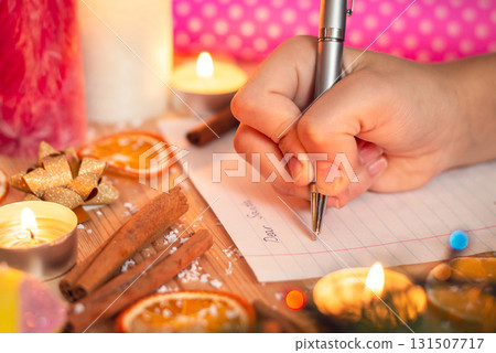 Close-up shot of hand of a child, writing a letter to Santa. Christmas decoration around, festive atmosphere, belief in magic and miracle. 131507717