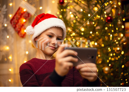 Cute young boy makes a selfie in the hat of Santa Claus, decorated fir-tree full of lights on the background. The concept of Christmas, holidays. Cute young boy makes a selfie in the hat of Santa Claus, decorated fir-tree full of lights on the background. The concept of Christmas, holidays. 131507723