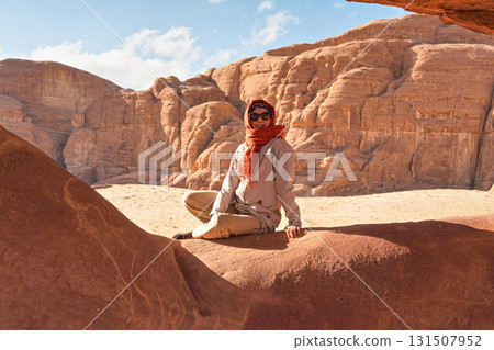 Young woman wearing warm jacket and scarf around head smiling, sitting at naturally formed rock window in wadi rum desert on sunny day 131507952