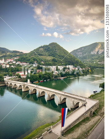 An old stone bridge with arches spans the tranquil Drina River, surrounded by green hills 131508098
