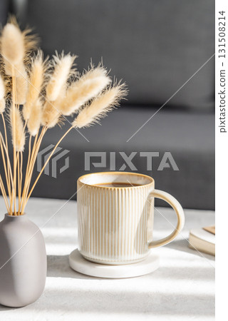 Beige cup of coffee on a light table with soft shadows, vase and notebook on the background 131508214