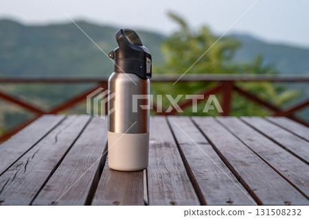 Stainless steel water bottle with black lid on rustic wooden table with nature and mountains  131508232