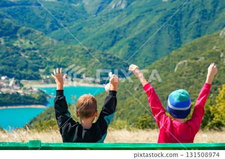 Close-up of two boys sitting on wooden bench with raised hands, looking at scenic Piva Lake and mountains in Montenegro. Concept of childhood, freedom and outdoor travel. 131508974