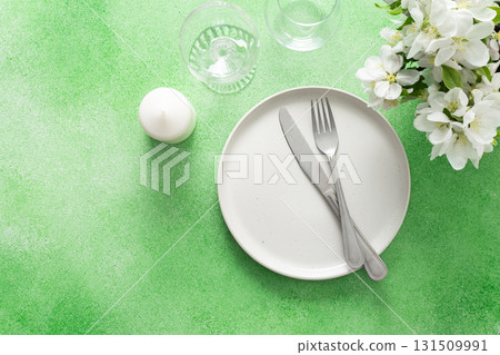 A lovely spring table setting features a white plate, silverware, and elegant glassware, complemented by fresh flowers and a soft green background, perfect for gatherings and meals A lovely spring table setting features a white plate, silverware, and elegant glassware, complemented by fresh flowers and a soft green background, perfect for gatherings and meals 131509991
