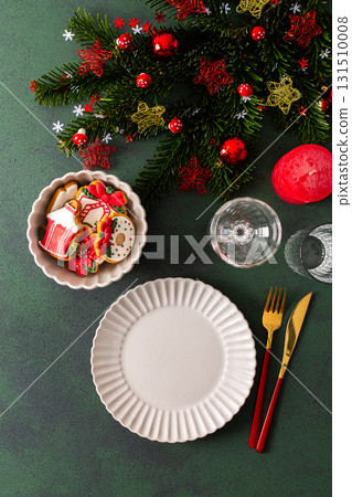 A Christmas table setting featuring an empty plate, napkin and gingerbread ,cutlery and a candle on a deep green background. Spruse branches and red tree decorations create a festive atmosphere 131510008