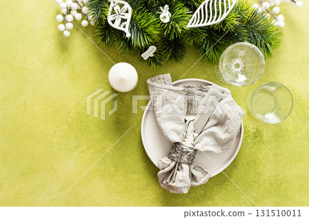 A top view of a Christmas table setting featuring an empty plate with napkin and cutlery, festive spruse and white tree decorations on a bright green background 131510011
