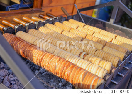 Traditional trdelnik pastries are roasting on rotating wooden spits over hot coals. Concept of trdelnik as a sweet street food, concept of trdelnik as cultural dessert 131510298