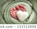 The tiny foot of a newborn baby. Soft feet of a new born in a olive green wool blanket. Close up of toes, heels and feet of a newborn. Knitted white heart in the legs of a baby. Macro photography. 131512608