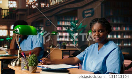 African american med school pupil studies pharmacology notes in a library, preparing for exams and advancing their healthcare career. Girl in scrubs works hard on medical school success. Camera A. 131513104