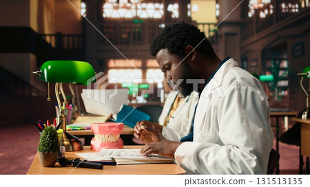 African american future dentist examines a plastic model of human teeth, studying stomatology practices in the campus library. Male student learns about dentistry or oral health. Camera A. African american future dentist examines a plastic model of human teeth, studying stomatology practices in the campus library. Male student learns about dentistry or oral health. Camera A. 131513135