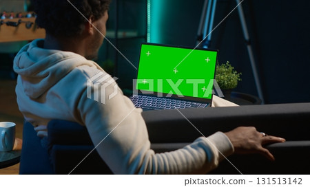 Happy pleased young man employs isolated copy space at home, relaxing on his couch in a modern living room decorated with neon lights and trendy furniture. Male person with gadgets. Camera A. 131513142