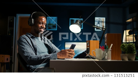Black remote worker takes a break to scroll on his mobile phone apps, texting messages and chatting with his friends online. Young self employed guy enjoys multitasking, listens to music. Camera A. Black remote worker takes a break to scroll on his mobile phone apps, texting messages and chatting with his friends online. Young self employed guy enjoys multitasking, listens to music. Camera A. 131513251