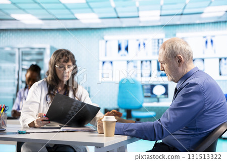 Female doctor evaluating x ray imaging test findings to provide the diagnostic report for a sick old man, meeting in her cabinet. Healthcare provider ensuring recovery plan on insurance. Female doctor evaluating x ray imaging test findings to provide the diagnostic report for a sick old man, meeting in her cabinet. Healthcare provider ensuring recovery plan on insurance. 131513322