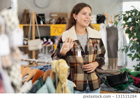 During November sales, young woman choose and try on checkered coat-jacket, view reflection in mirror. During November sales, young woman choose and try on checkered coat-jacket, view reflection in mirror. 131515284