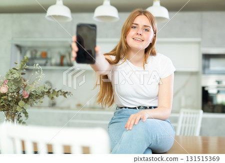 Portrait of cheerful teen girl showing modern mobile phone with black screen and sitting on kitchen countertop. Gadget with empty free space for mock up Portrait of cheerful teen girl showing modern mobile phone with black screen and sitting on kitchen countertop. Gadget with empty free space for mock up 131515369