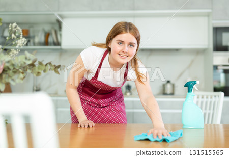 Smiling teenage girl in apron using spray and wiping dust on countertop with rag while cleaning modern kitchen Smiling teenage girl in apron using spray and wiping dust on countertop with rag while cleaning modern kitchen 131515565