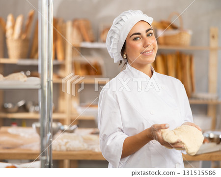 Young female baker stands in bakery holding a piece of dough Young female baker stands in bakery holding a piece of dough 131515586