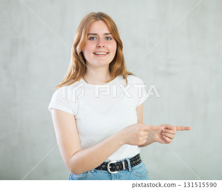 Portrait of cheerful charismatic teen girl in white t-shirt and blue jeans pointing to right against gray wall with copy space Portrait of cheerful charismatic teen girl in white t-shirt and blue jeans pointing to right against gray wall with copy space 131515590