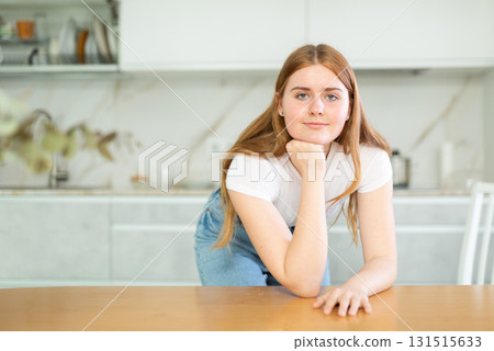 In kitchen with light furniture, long-haired girl put elbows on table and props chin with fist In kitchen with light furniture, long-haired girl put elbows on table and props chin with fist 131515633