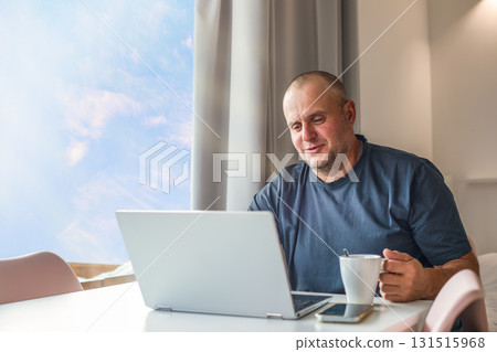 Middle-aged man drinking morning coffee and communicating via laptop. 131515968