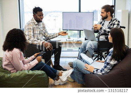 Team of three young adults and one mid adult discussing project in modern workspace. Diverse group with wheelchair user and mix of ethnicities fostering inclusion. Team of three young adults and one mid adult discussing project in modern workspace. Diverse group with wheelchair user and mix of ethnicities fostering inclusion. 131518217