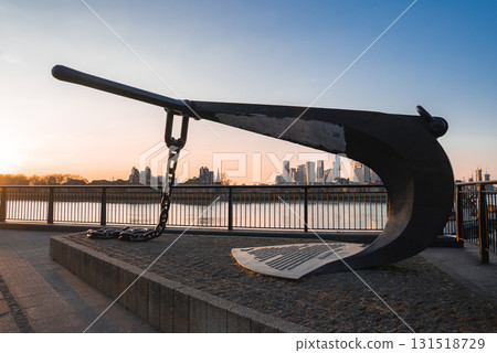 A large black anchor sculpture on a raised platform near the Thames River, with Canary Wharf's modern skyscrapers glowing in a warm sunset light. 131518729