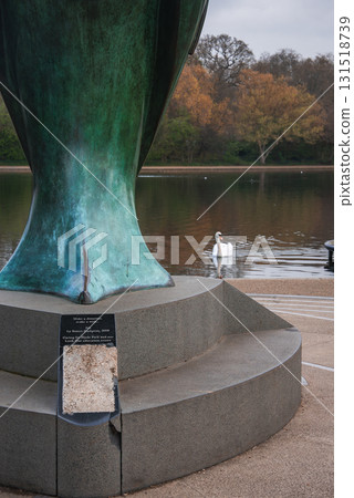 A green patinated sculpture on a stone pedestal with a plaque near a calm body of water, a white swan, and autumn colored trees in a London park. 131518739