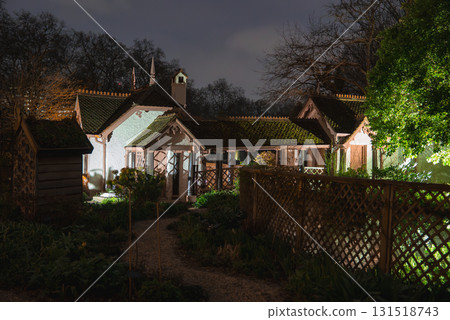 A historic cottage in London illuminated at night, featuring steep roofs, decorative trim, a chimney, and a lush garden with wooden lattice fencing. 131518743